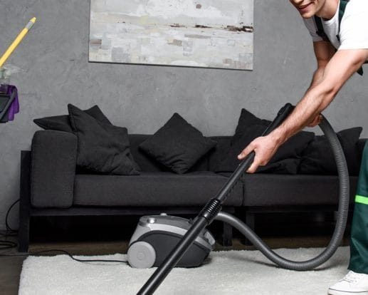 Individual using a vacuum cleaner on a sofa in a living room setting with grey walls.
