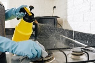 Individual using a yellow spray bottle to clean a kitchen stove with a cloth.